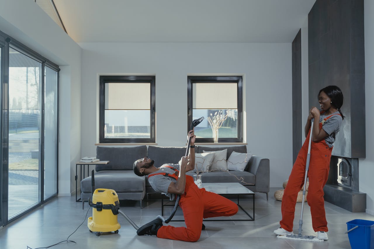 services-03 Fun and engaging cleaning session with a man and woman in overalls using cleaning tools in a modern living room.