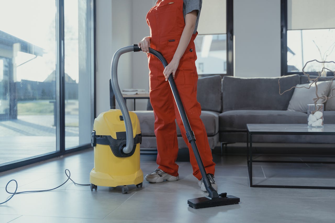 gallery-6 A professional cleaner in red coveralls vacuuming a modern living room with a yellow vacuum cleaner.