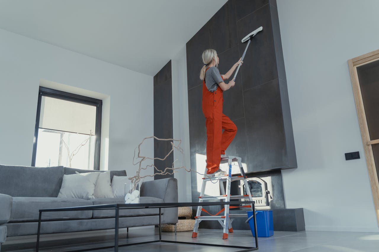 hero-img-01 A woman stands on a ladder in a modern living room, cleaning the high wall with a mop.